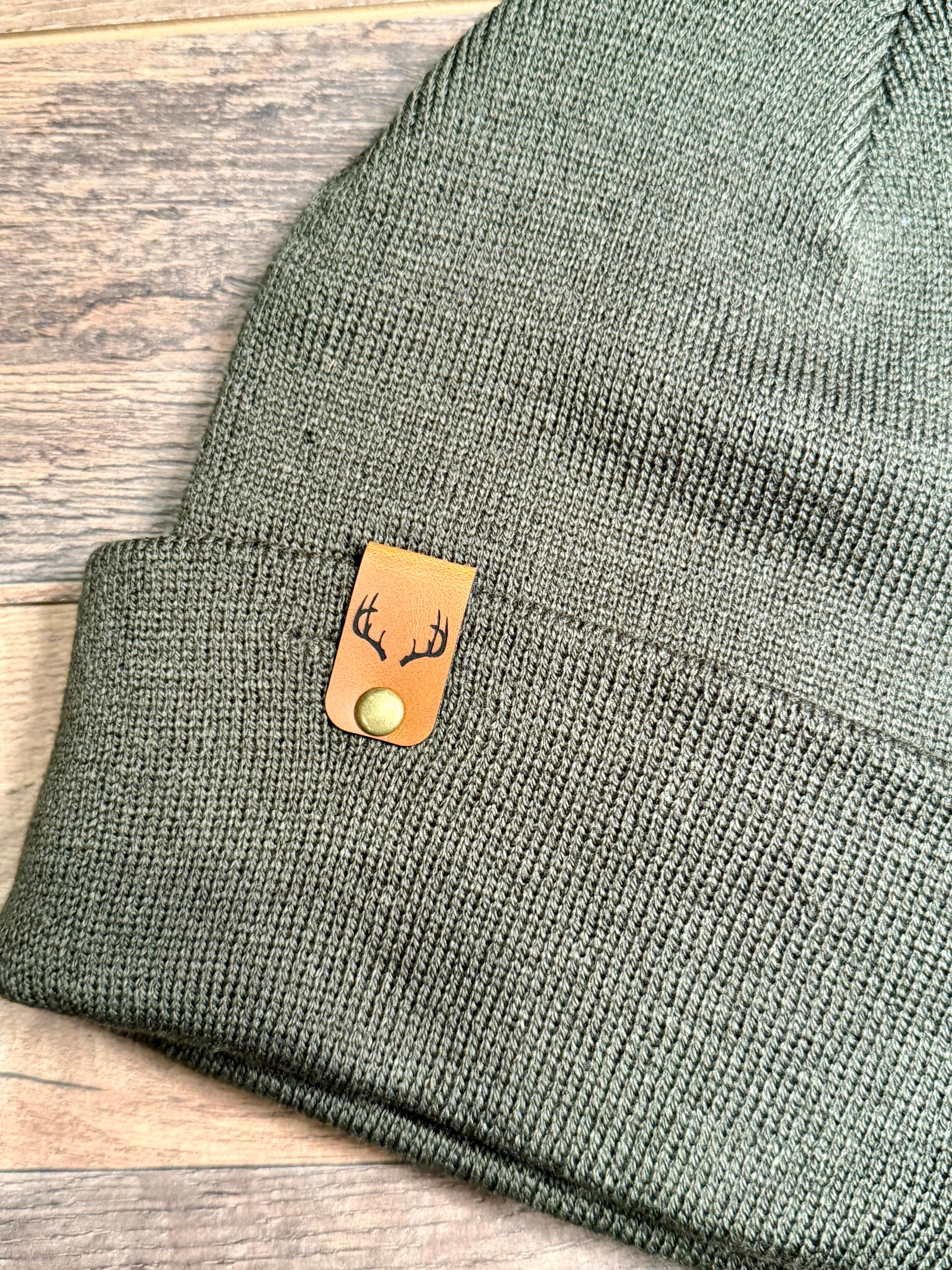 Green knit beanie with a leather tag featuring deer antlers on a wooden surface