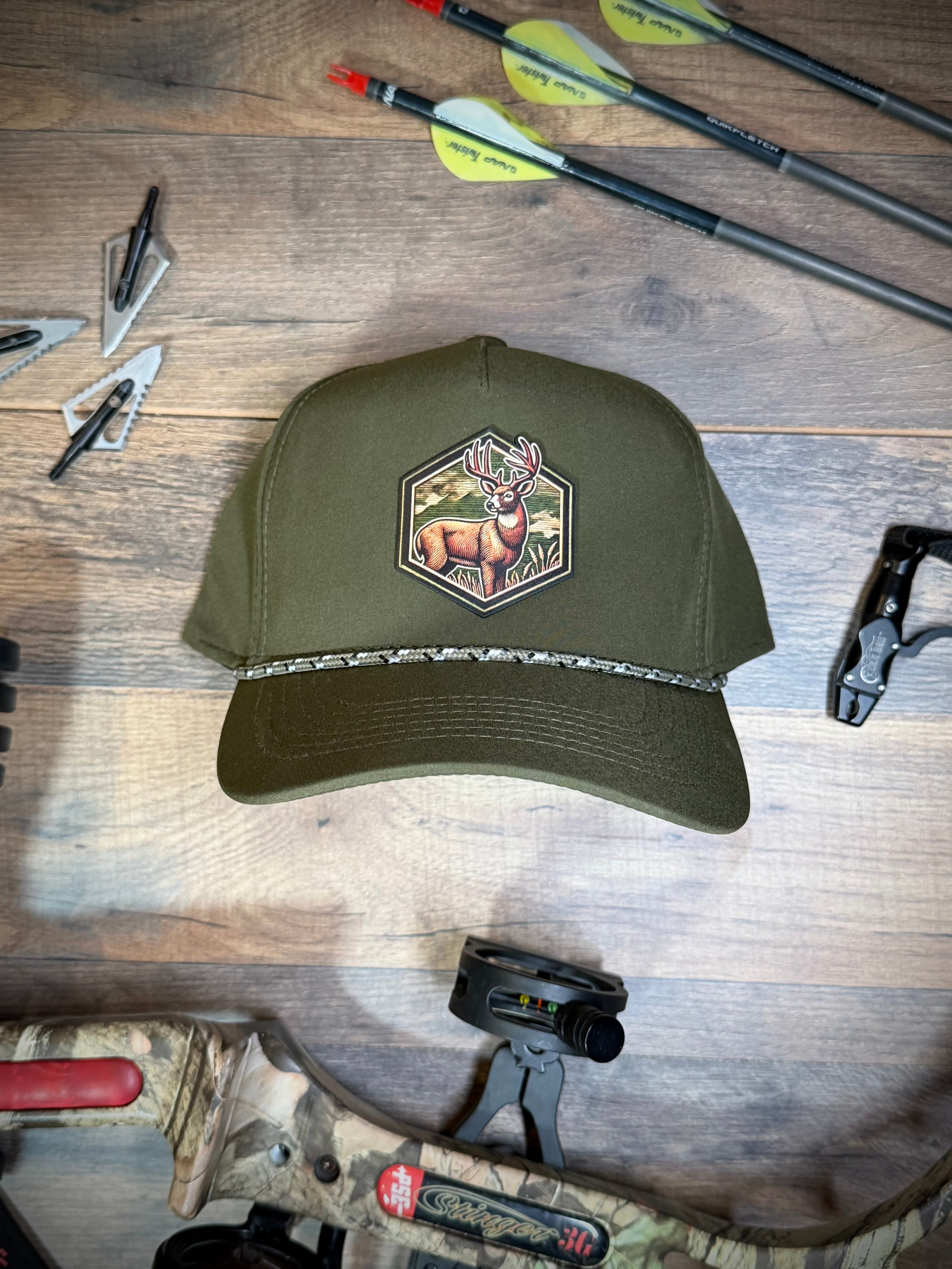 Green hunting cap with a deer emblem on a wooden surface with arrows and a rifle.