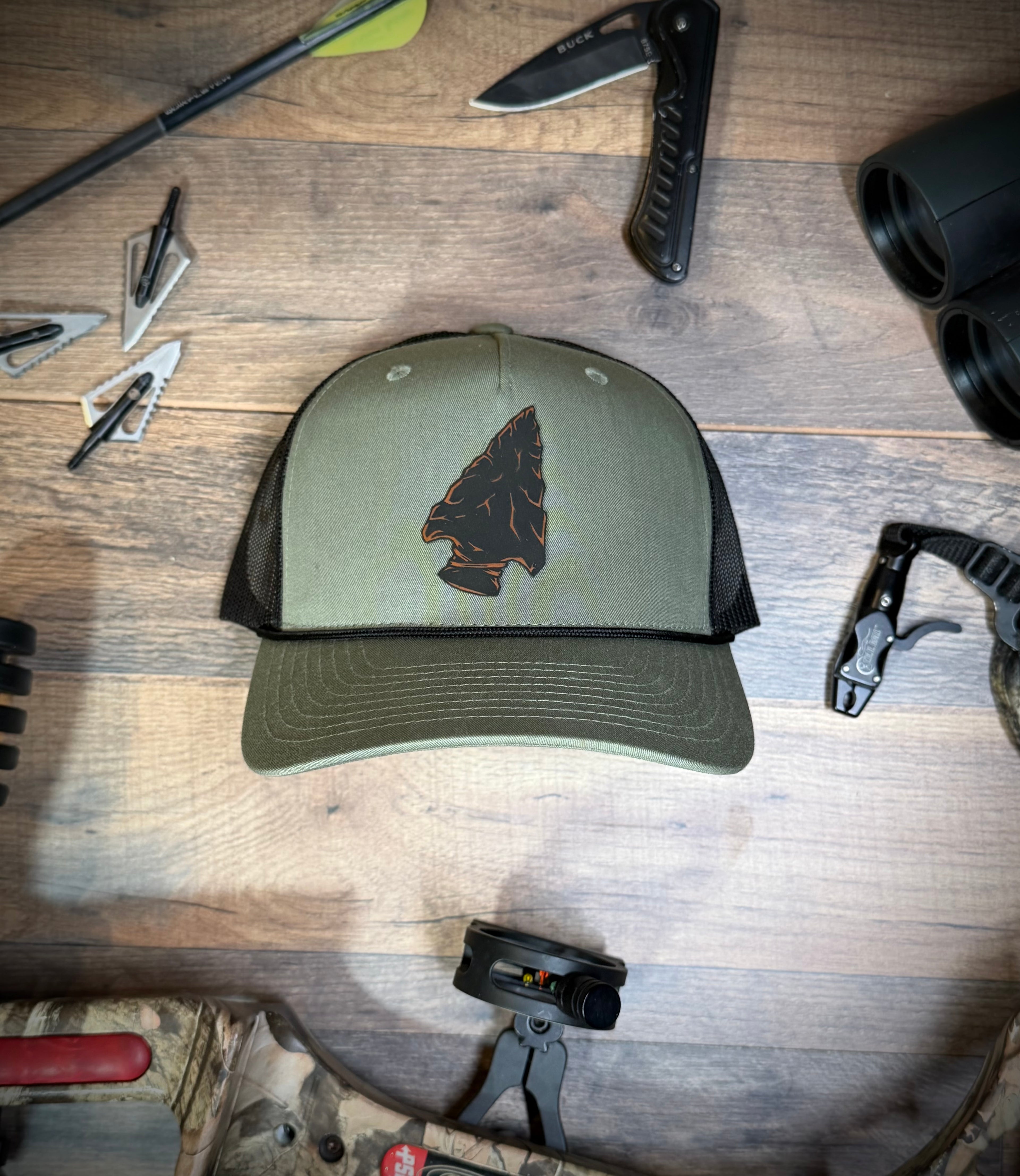 Green cap with an arrowhead design on a wooden surface with hunting gear.