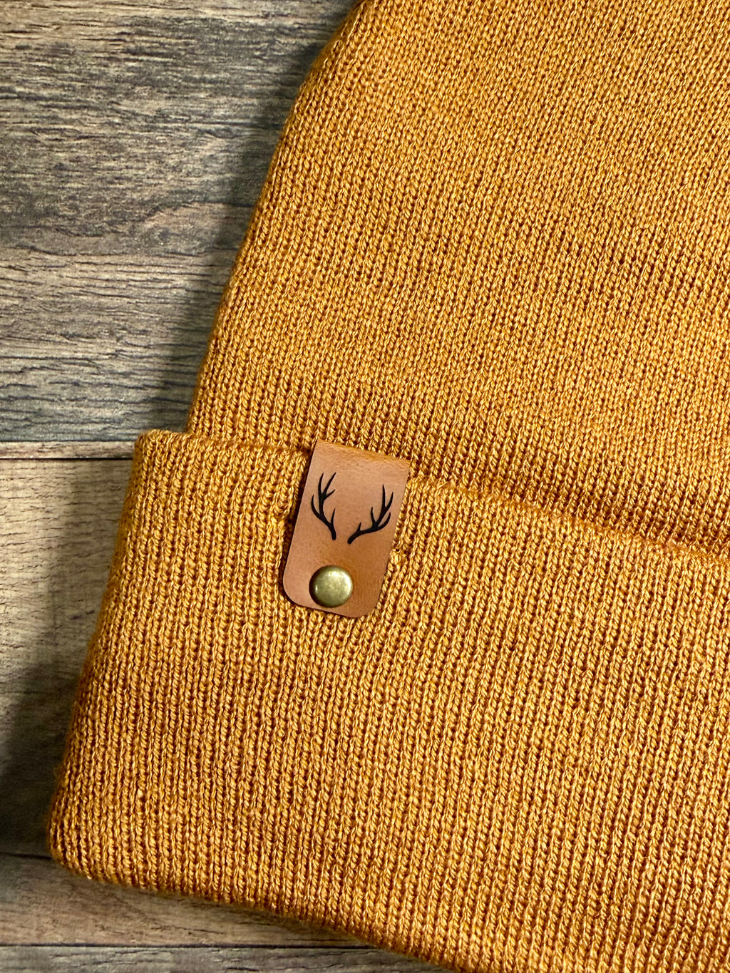 Brown knit beanie with a leather patch featuring deer antlers on a wooden surface