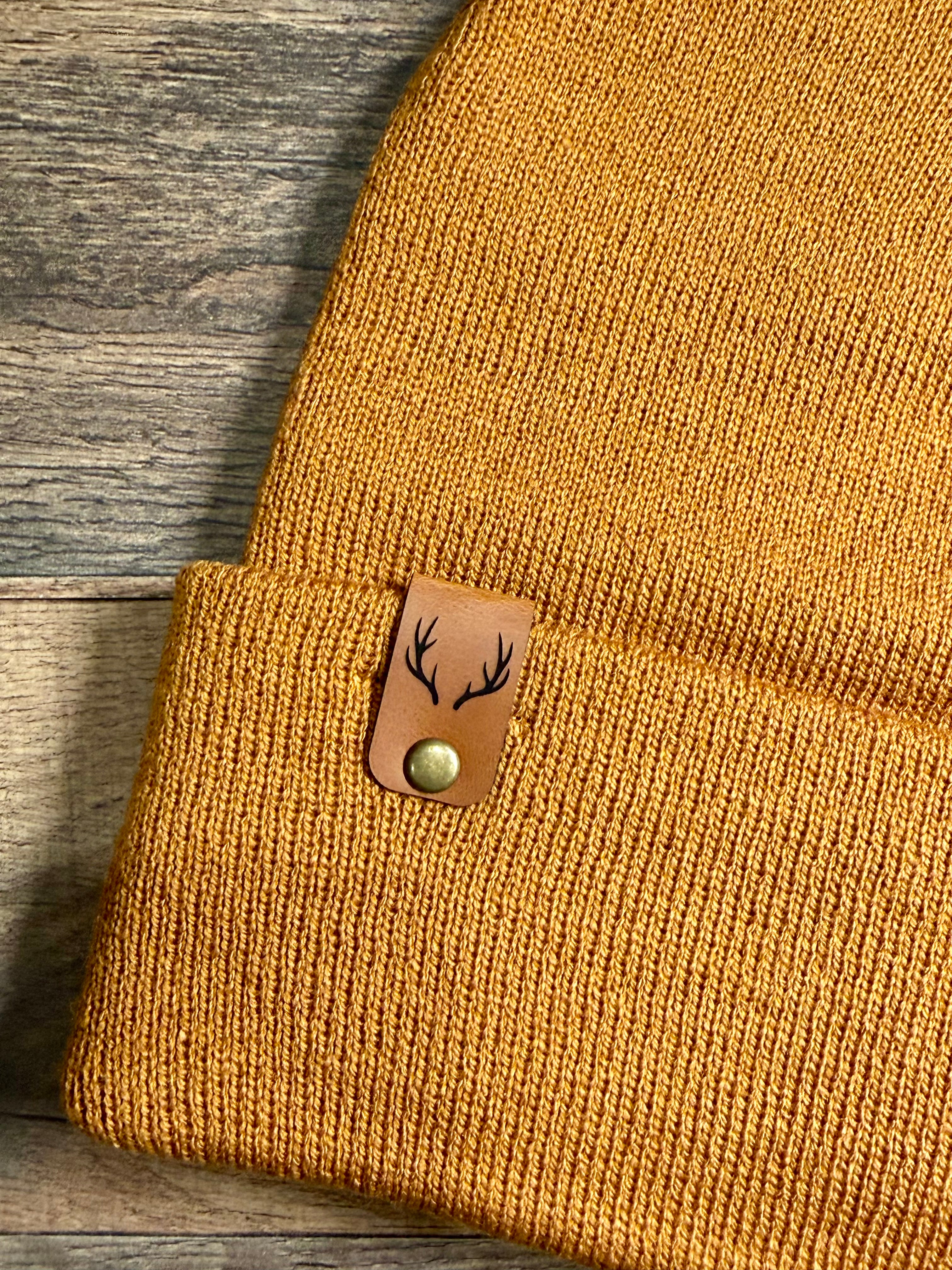 Brown knit beanie with a leather patch featuring deer antlers on a wooden surface