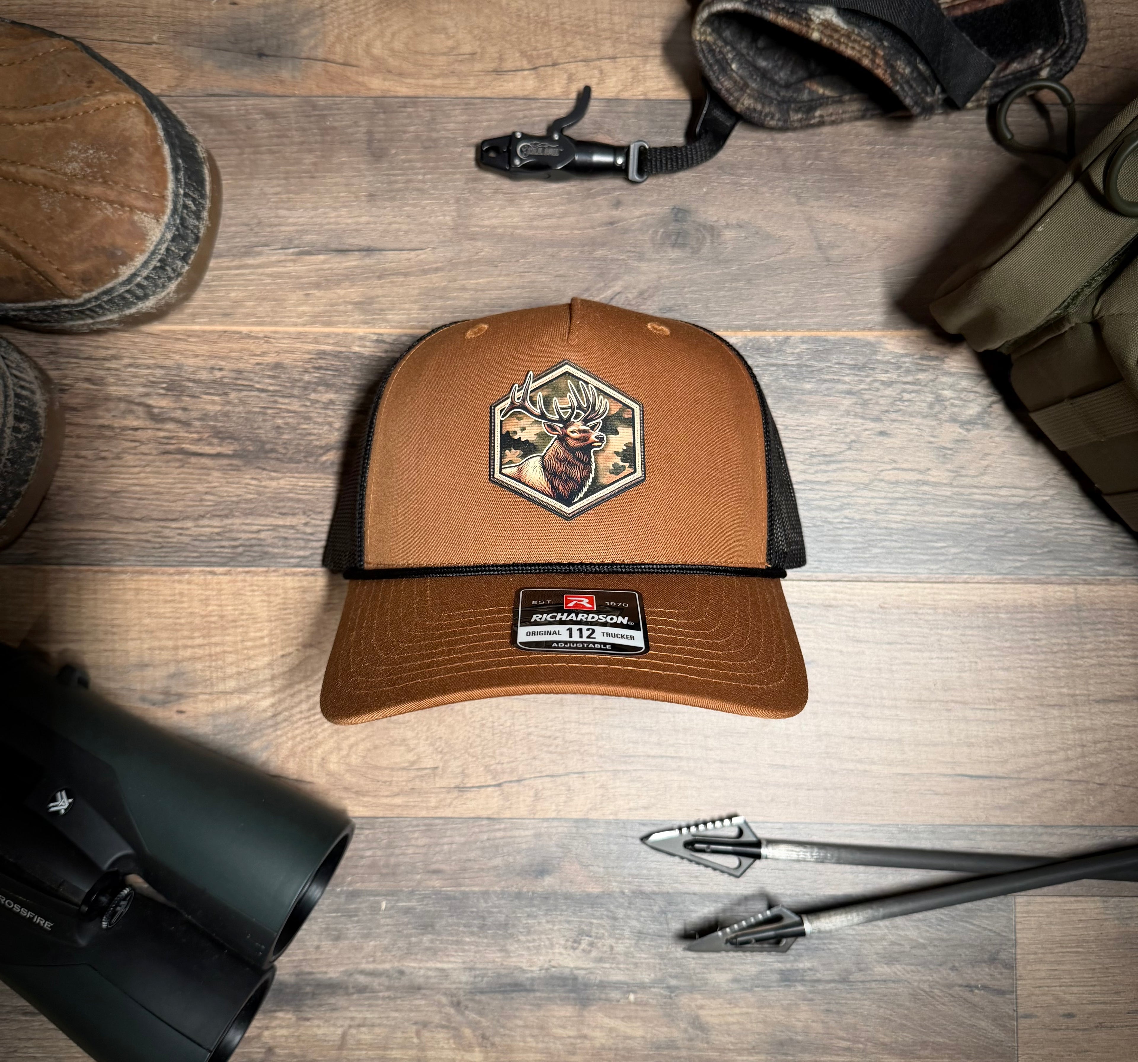 Brown cap with hunting design on a wooden surface with binoculars and a bow.
