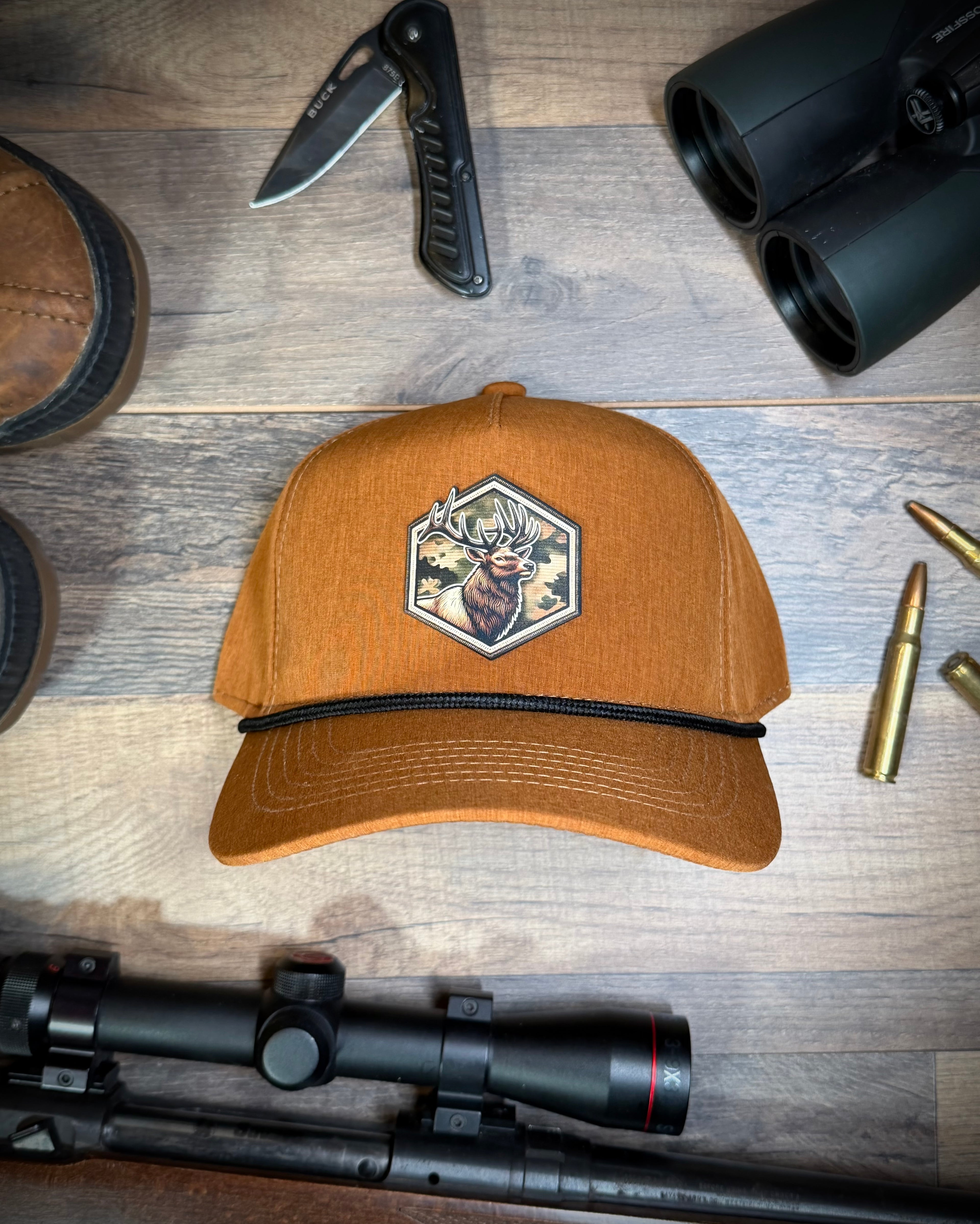 Brown cap with a logo on a wooden surface with hunting gear around