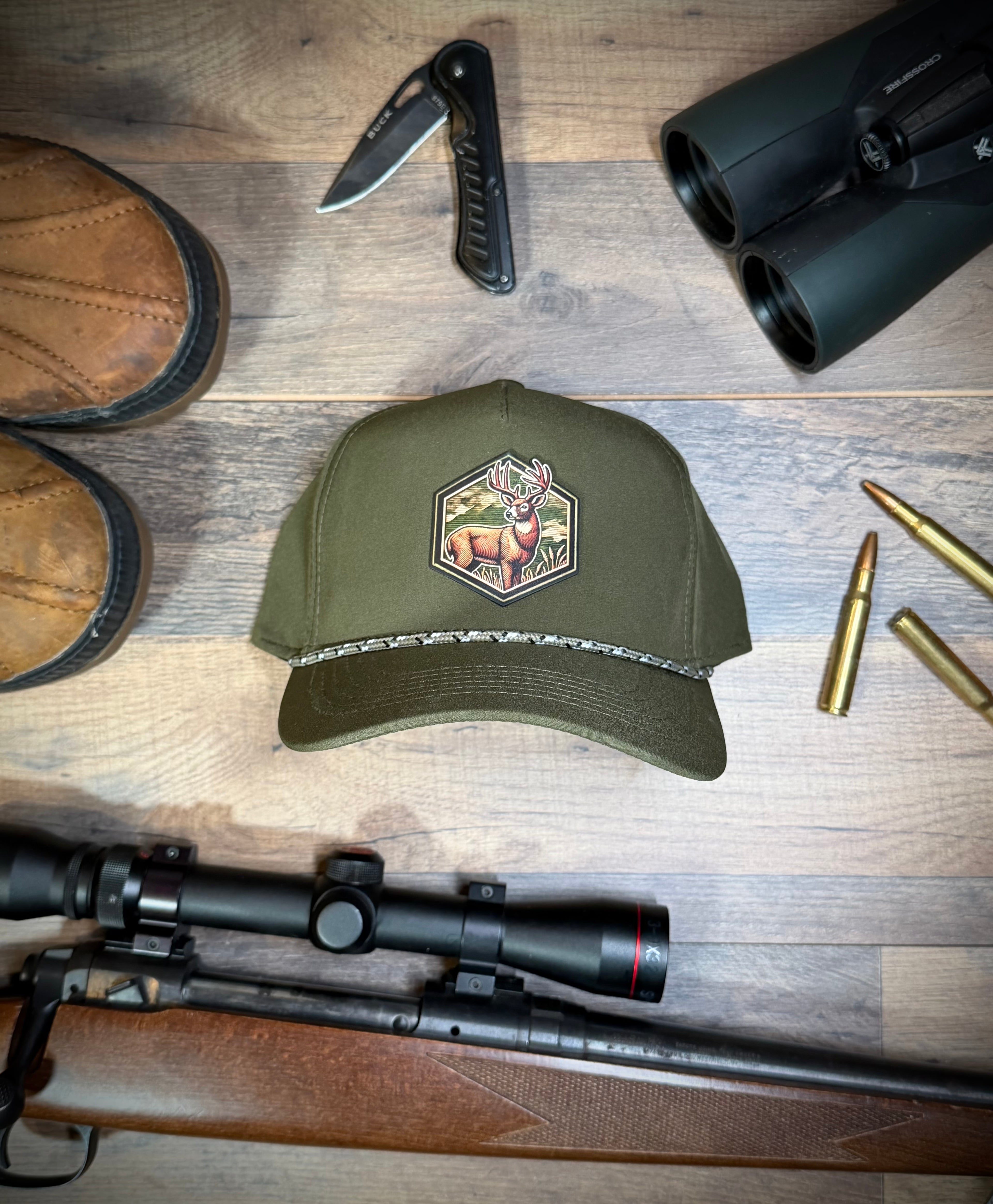 Green hunting cap with a deer emblem on a wooden surface with binoculars, rifle, boots, and bullets.