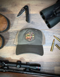 Gray cap with a deer emblem on a wooden surface with hunting gear.